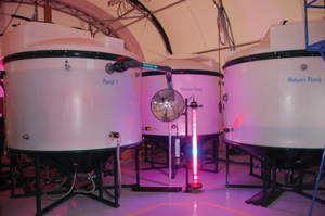 The demonstration plant at St Marys Cement, showing the company’s proprietary high intensity illuminator in the centre.  The tank on the left is a bioreactor, and there is a second bioreactor to its left out of frame.  Both of these bioreactors overflow into the harvest tank in the centre. From there the algae is sent to the separator, which is out of frame to the right and the recovered water is then recycled back to the return tank (on the right).  Finally, the water is returned to the bioreactor and the loop is closed.